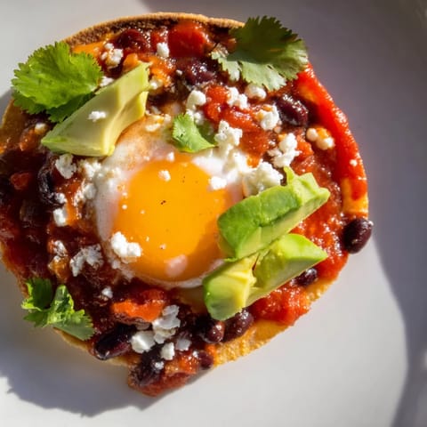A close-up view of Huevos Rancheros on a rustic plate, featuring crumbled queso fresco, black beans, and lime wedges ready for squeezing.