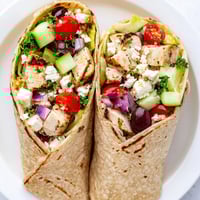 Golden-brown grilled chicken pieces atop vibrant Greek salad with crumbled feta, tucked inside a warm flour tortilla.  